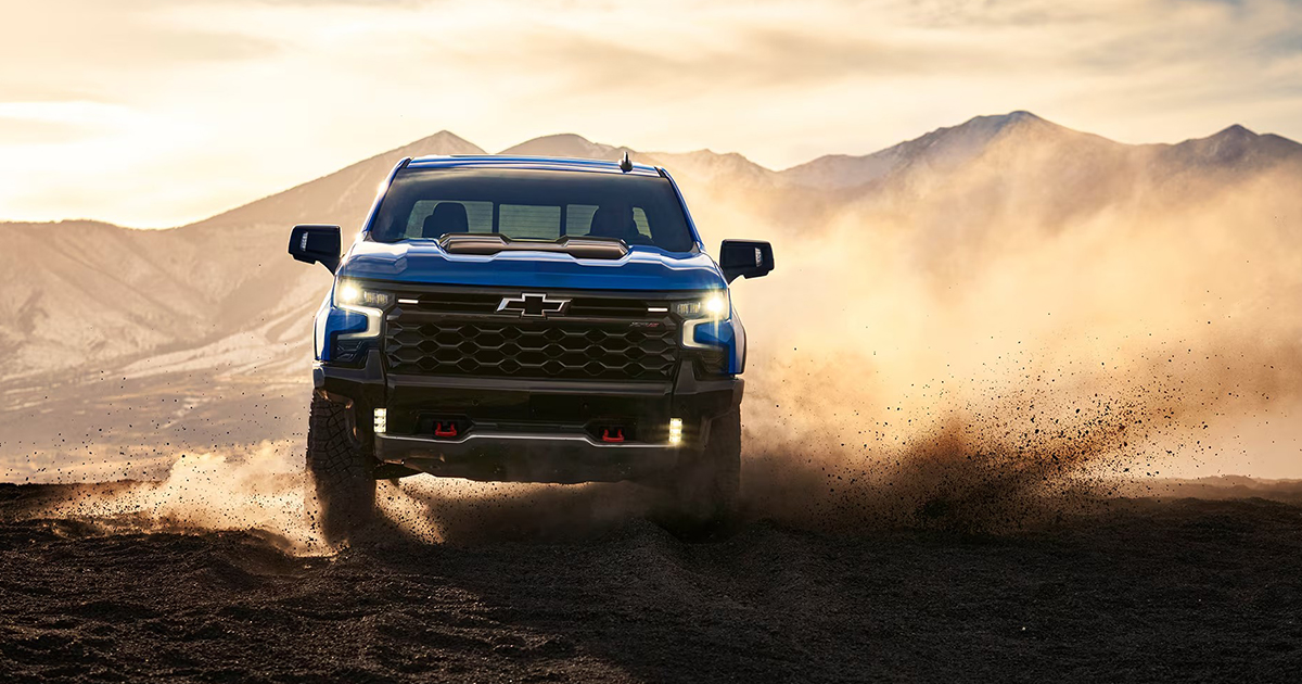 Blue 2025 Chevrolet Silverado 2500HD or 3500HD with hood scoop jumping through desert sand with mountains in background
