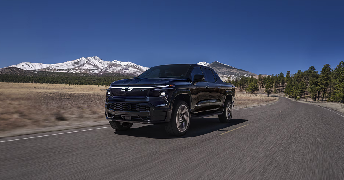 Black Chevrolet Silverado EV driving on mountain highway with snow-capped peaks in background