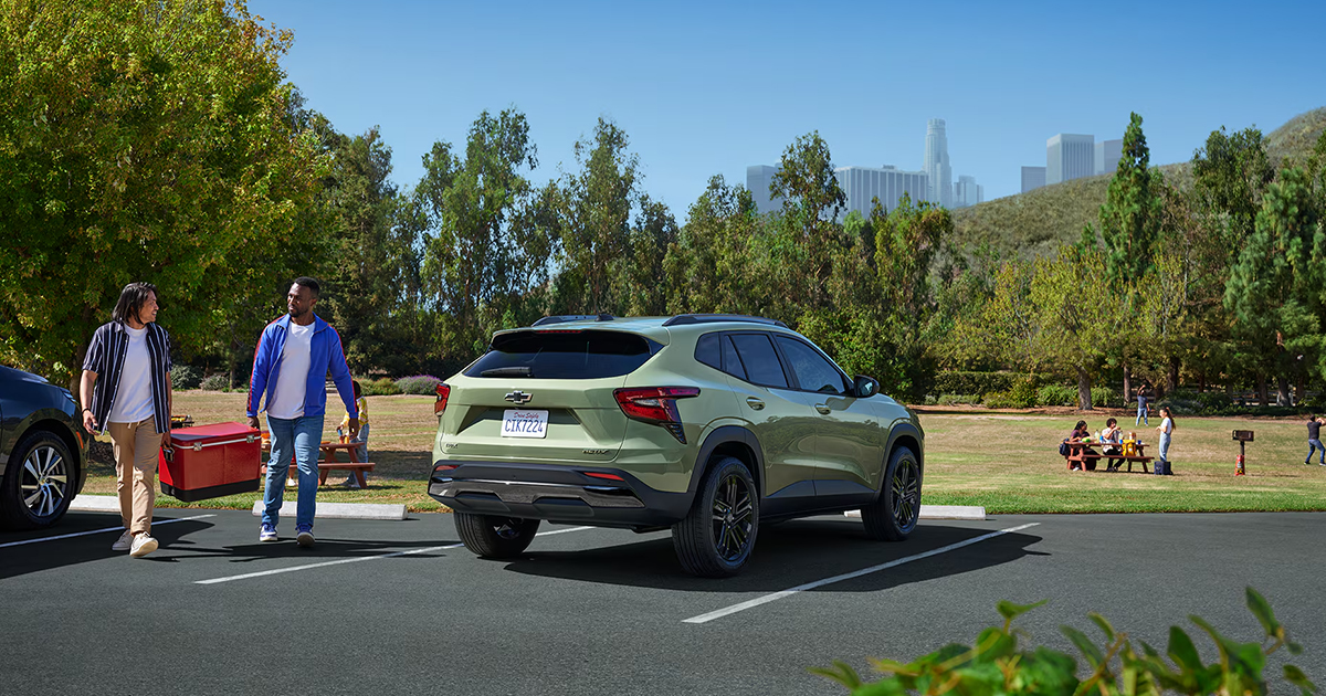 Light green 2025 Chevrolet Trax Activ at park with two people carrying cooler and city skyline in background