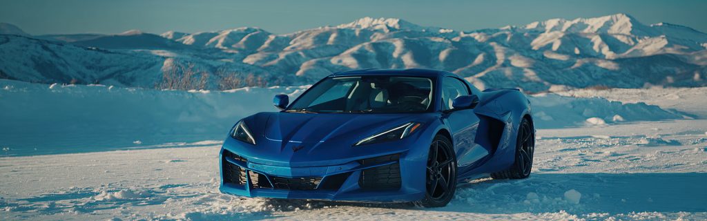A blue Chevrolet Corvette sports car parked on a snow-covered field with sunlit, snow-capped mountains in the background.