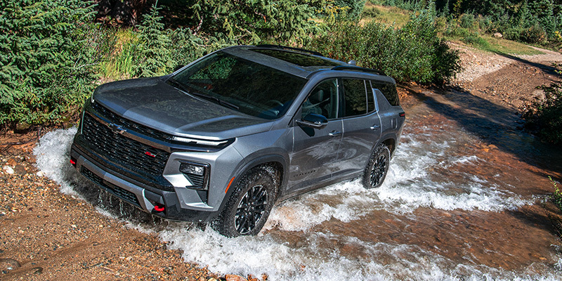 A grey Chevrolet Traverse Z71 SUV driving through a shallow, rushing stream in a forested area.