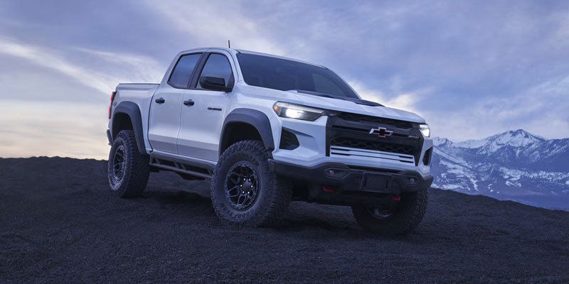 A white Chevrolet Colorado ZR2 pickup truck parked on a dark, rocky hill with mountains in the background under a cloudy twilight sky.