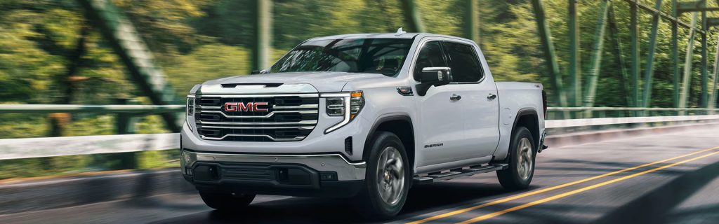 White GMC Sierra 1500 pickup truck driving across a steel bridge surrounded by lush greenery.