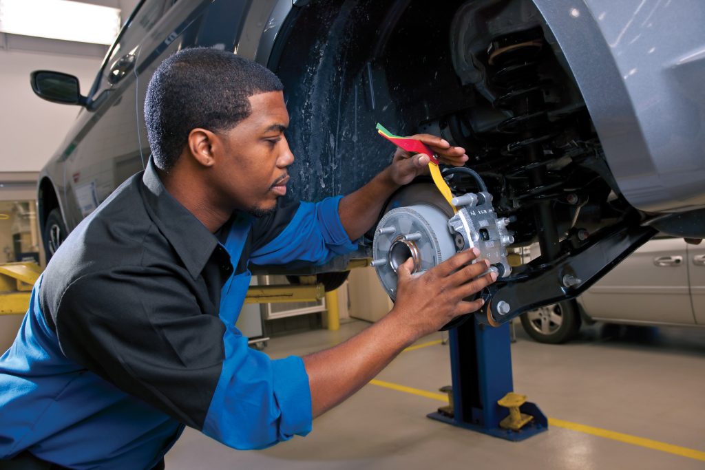 Technician inspecting a vehicle brake assembly while wearing safety glasses and gloves in a service bay.