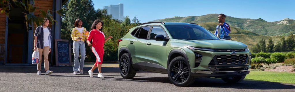 A group of friends walking past a green Chevrolet Trax crossover SUV parked outside a market with hills in the background.